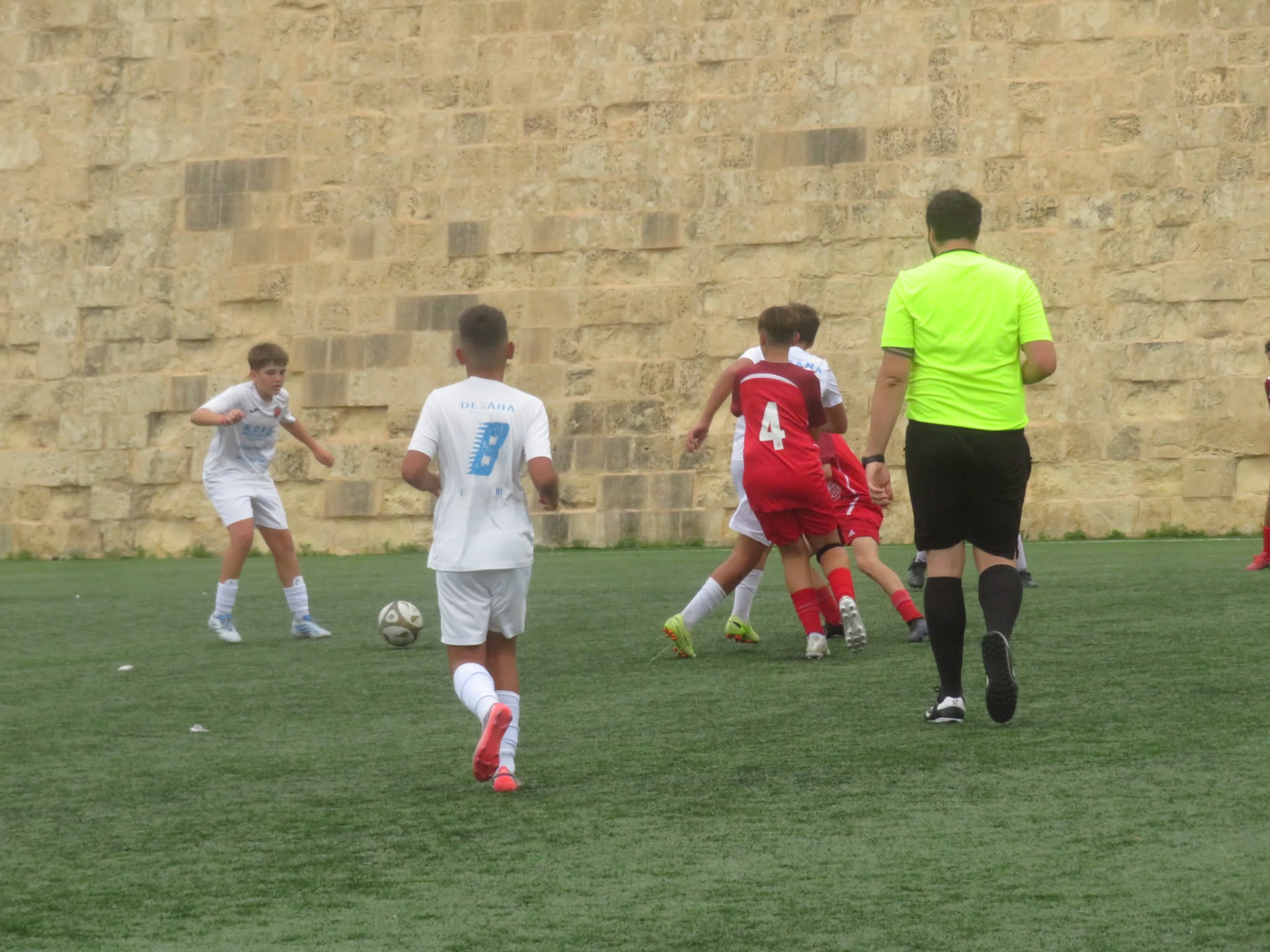 Plejers żgħażagħ bil-flokk aħmar u abjad jiġru wara l-ballun fuq grawnd sintetiku quddiem ħajt tal-ġebla, b'referee bl-isfar.